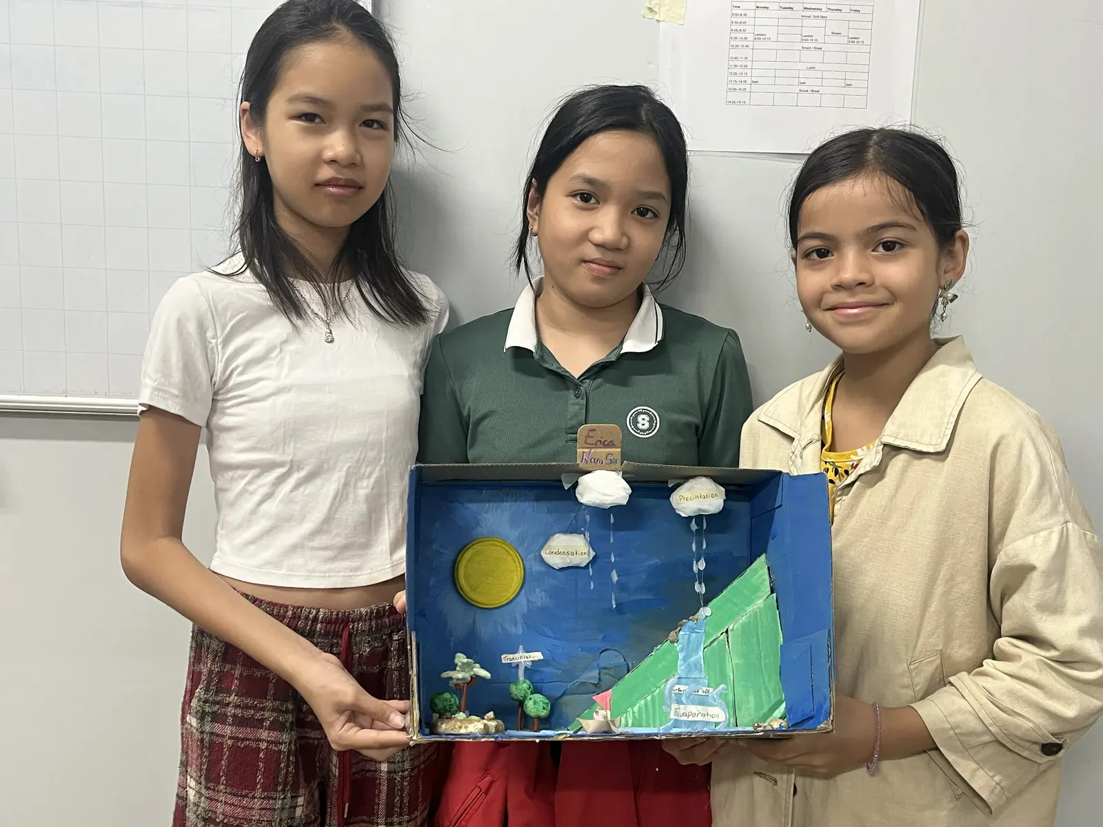 Three students hold up their water cycle model, built in a box painted blue, with a paper sun and cotton-wool clouds labelled condensation and precipitation