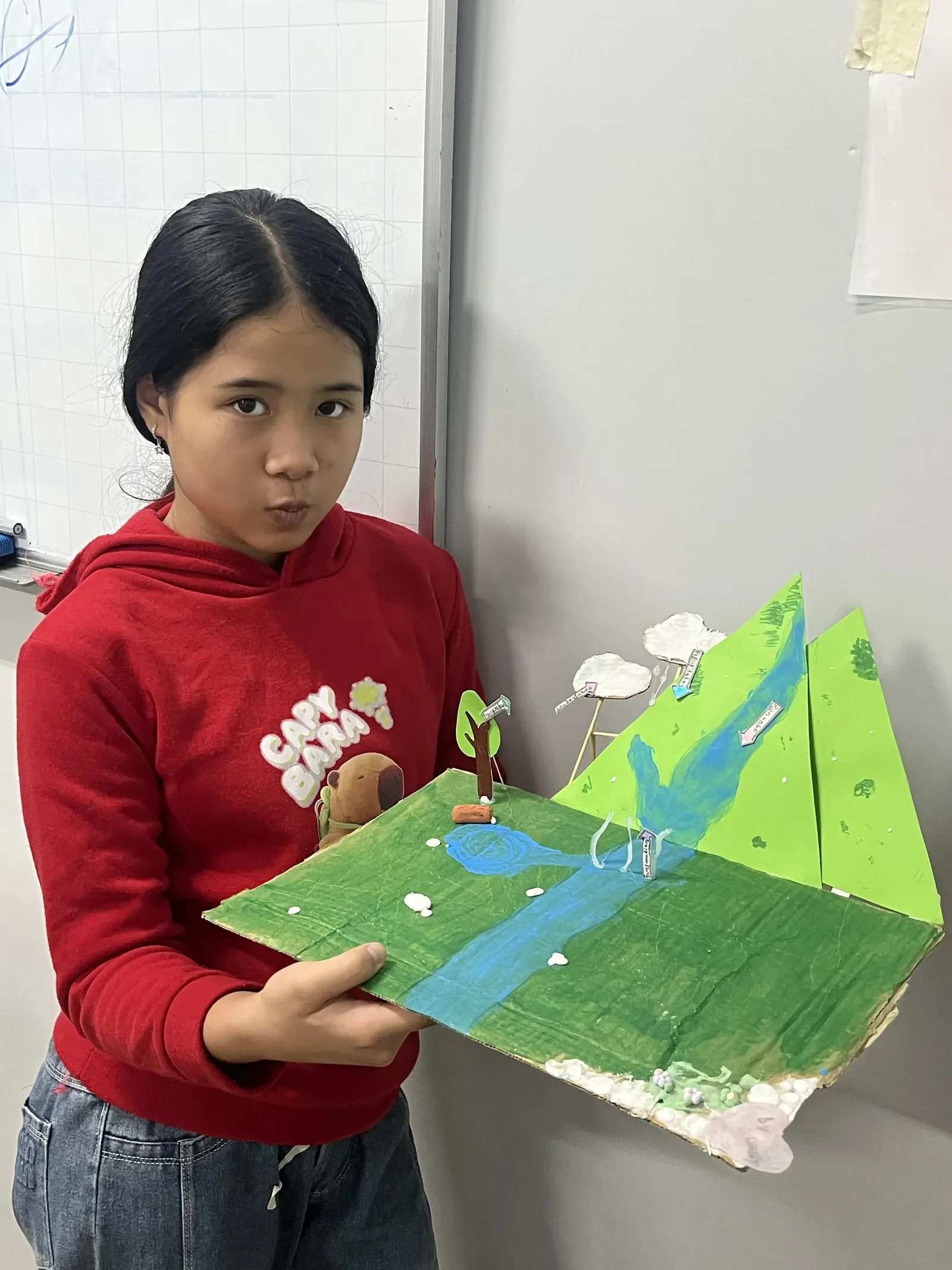 A student holds up her water cycle model: a painted river running down green card hills, with clouds on sticks and labelled arrows