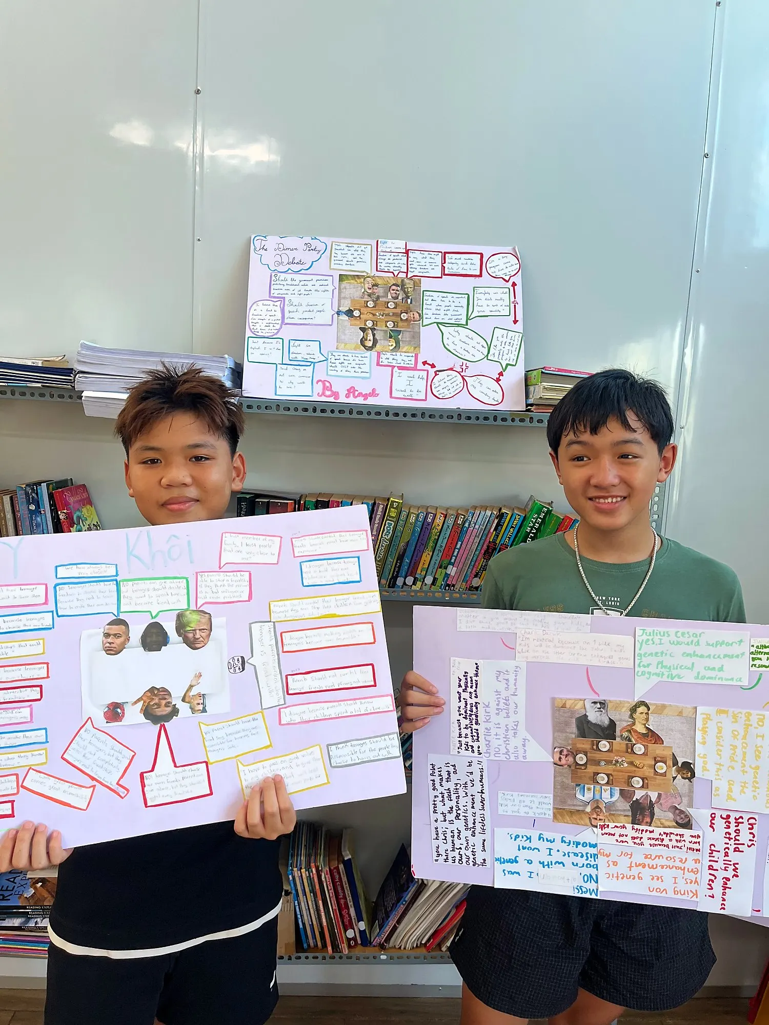 Hai học viên Upper Secondary cầm poster bữa tiệc tranh luận của mình trong thư viện