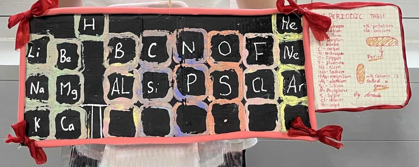 Amada's finished periodic table: the first 20 elements painted on black squares in a pink clay frame with red bows, and her handwritten key beside it